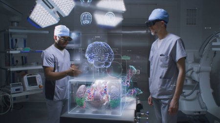Foto de Diversos cirujanos masculinos en auriculares AR trabajan en quirófano usando la exhibición holográfica. Gráficos 3D de esqueleto humano virtual, órganos y signos vitales. Tecnología de IA en medicina. Asistencia sanitaria futurista. - Imagen libre de derechos