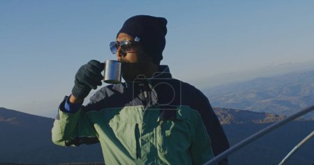 Foto de Hombre afroamericano en gafas de sol se encuentra cerca de la tienda y mira el hermoso valle del bosque. Turista bebe taza de té caliente y se calienta en la cima de la colina de la montaña. Mochilero se detuvo a descansar durante una caminata. - Imagen libre de derechos