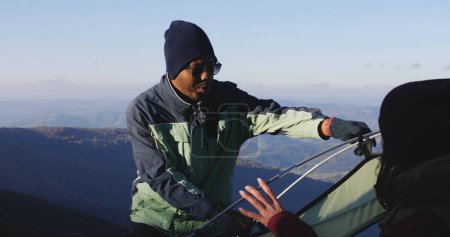Photo for African American man sets up tent on top of mountain hill. Woman gives cup of tea to backpacker friend. Multiethnic family on adventure trip. Diverse tourist couple stopped to rest during a hike. - Royalty Free Image