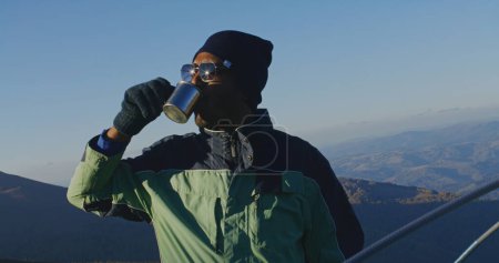 Photo for African American man in sunglasses stands near tent and looks at beautiful forest valley. Tourist drinks cup of hot tea and warms up on top of mountain hill. Backpacker stopped to rest during a hike. - Royalty Free Image