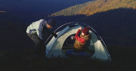 Téléchargez les photos : L'homme afro-américain installe une tente au sommet d'une colline à l'aide d'une hache. Femme boit une tasse de thé et se réchauffe dans la tente. Famille multiethnique en voyage d'aventure. Couple touristique arrêté pour se reposer pendant une randonnée. - en image libre de droit