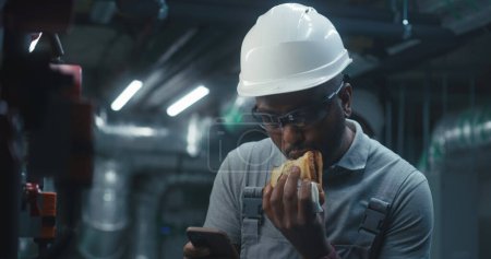 Photo for African American engineer eats sandwich and surfs the Internet on phone. Professional worker in uniform and protective hard hat having break working on industrial factory or energy facility. Close up. - Royalty Free Image