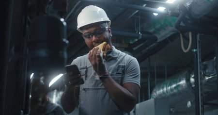 Photo for African American technician eats sandwich and surfs the Internet on mobile phone. Professional engineer in safety uniform and protective hard hat having break working on modern industrial factory. - Royalty Free Image