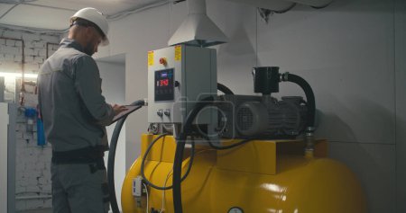 Professional engineer in safety uniform, goggles and protective hard hat adjusts vacuum pump station or air compressor using tablet computer. Male employee works on modern plant or industrial factory.
