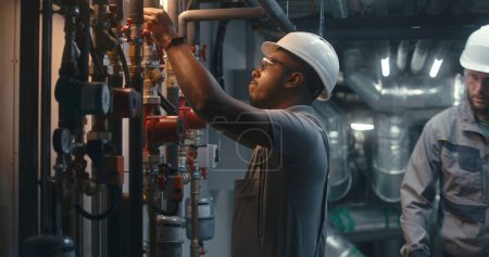 Photo for African American heavy industry worker wearing safety uniform and hard hat inspects piping system on modern plant or industrial energy facility. Professional engineer maintains manufacturing factory. - Royalty Free Image