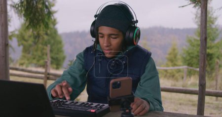 Photo for Young musician in headphones plays MIDI controller outdoors. African American teenager creates and records music sitting in wooden gazebo in mountains, using laptop and phone on tripod. Slow motion. - Royalty Free Image