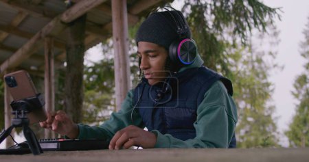 Téléchargez les photos : Adolescent afro-américain dans les écouteurs joue sur le clavier MIDI à l'extérieur, se déplace rythmiquement. Le jeune musicien crée et enregistre de la musique assis dans un kiosque en bois, en utilisant le téléphone sur le trépied. Mouvement lent - en image libre de droit