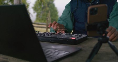 Téléchargez les photos : Musicien afro-américain joue sur contrôleur MIDI à l'extérieur. Jeune compositeur crée et enregistre de la musique, maîtrise de nouvelles chansons tout en étant assis dans le belvédère, en utilisant un ordinateur portable et un téléphone sur trépied. Ferme là. Mouvement lent - en image libre de droit