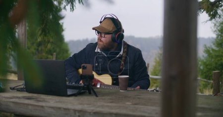 Photo for Caucasian man in headphones plays guitar and records music sitting in wooden gazebo. Professional male musician, composer masters new song outdoors in the forest. Music creating and production. - Royalty Free Image