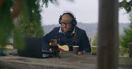 Photo for Caucasian man in headphones plays guitar sitting in wooden gazebo in the forest. Male musician composes and records music using laptop and phone on tripod during vacation trip in the mountains. - Royalty Free Image