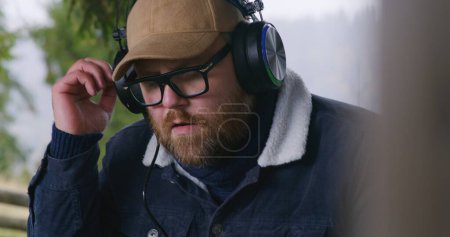 Photo for Close up of male musician in glasses and headphones playing MIDI keyboard, composing music and moving rhythmically sitting outdoors. Caucasian man records new song during vacation to mountains. - Royalty Free Image