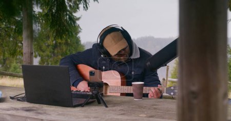 Photo for Caucasian man in headphones sings, plays guitar in wooden gazebo in the mountains. Male musician creates and records music or song outdoors using laptop, phone on tripod and professional microphone. - Royalty Free Image
