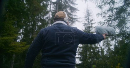 Téléchargez les photos : Le concepteur sonore portant un casque enregistre les sons de la nature pour le film dans la forêt de conifères à l'aide d'un microphone à pare-brise en fourrure. Homme caucasien travaille avec un équipement audio professionnel à l'extérieur. Mouvement lent. - en image libre de droit