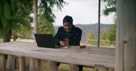 Photo for African American businessman sits in wooden gazebo, talks at online conference using laptop and phone on tripod. Traveler remotely consults clients during vacation in mountain forest. Outdoor working. - Royalty Free Image