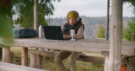 Téléchargez les photos : Le jeune garçon utilise un ordinateur portable et un téléphone sur trépied pour diffuser et jouer à des jeux pendant les vacances dans la forêt de montagne. Streamer est assis dans un kiosque en bois et communique avec les téléspectateurs à l'aide d'un microphone professionnel - en image libre de droit