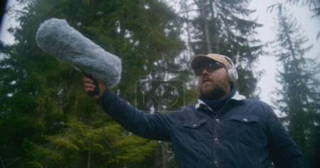 Téléchargez les photos : Un homme caucasien portant un casque enregistre les sons de la nature pour un film en bois de conifères à l'aide d'un microphone à pare-brise à fourrure. Ingénieur du son travaille avec des équipements audio professionnels à l'extérieur. Mouvement lent. - en image libre de droit