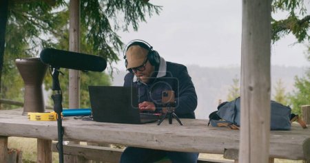 Téléchargez les photos : Musicien caucasien dans les écouteurs utilise le clavier MIDI pour composer la chanson assis dans le gazebo dans la forêt. Homme adulte crée et enregistre de la musique en utilisant le téléphone sur trépied et ordinateur portable pendant les vacances en montagne. - en image libre de droit