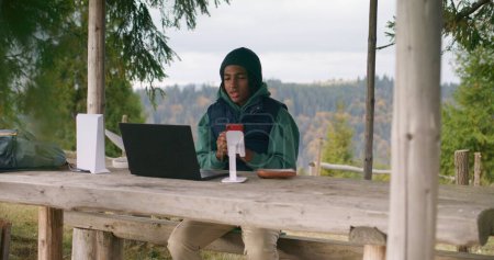 Téléchargez les photos : African American jeune garçon est assis dans un kiosque en bois et parle lors d'un appel vidéo à l'aide d'un ordinateur portable et d'un téléphone sur trépied. Adolescent étudie à distance pendant les vacances dans la forêt de montagne. Concept d'étude en plein air. - en image libre de droit