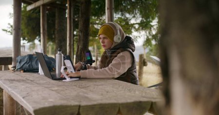 Photo for Teenager in headphones uses laptop and phone on tripod to create content for viewers during holidays in mountain forest. Young streamer sits in wooden gazebo and talks into professional microphone. - Royalty Free Image