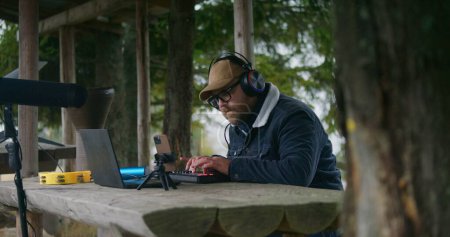 Photo for Male musician in headphones plays on MIDI keyboard, creates music and moves rhythmically sitting in gazebo in forest. Adult Caucasian man uses laptop and phone on tripod to record song outdoors. - Royalty Free Image