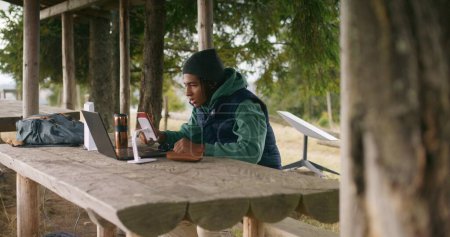 Téléchargez les photos : African American jeune garçon est assis dans un kiosque en bois, parle lors d'un appel vidéo à l'aide d'un ordinateur portable et téléphone sur trépied. Adolescent étudie à distance pendant les vacances dans la belle forêt de montagne. Concept d'étude extérieur. - en image libre de droit