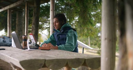 Téléchargez les photos : African American jeune garçon est assis dans un kiosque en bois, parle avec le professeur lors d'un appel vidéo à l'aide d'un ordinateur portable et téléphone sur trépied. Tourisme à distance étudie pendant les vacances dans la forêt de montagne. Concept d'étude extérieur. - en image libre de droit