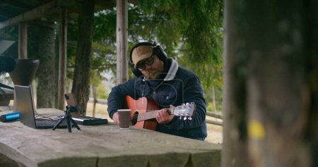 Téléchargez les photos : Homme caucasien dans les écouteurs joue de la guitare assis dans un kiosque en bois dans la forêt. Musicien professionnel crée et enregistre de la musique à l'aide d'un ordinateur portable et d'un téléphone sur trépied pendant le voyage de vacances dans les montagnes. - en image libre de droit