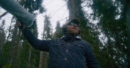 Photo for Caucasian man wearing headphones records sounds of nature for movie in coniferous wood using furry windscreen microphone. Sound engineer works with professional audio equipment outdoors. Low angle. - Royalty Free Image