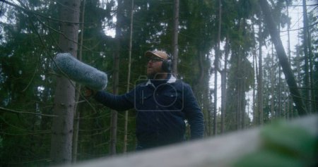Téléchargez les photos : Un homme caucasien portant un casque enregistre les sons de la nature pour un film en bois de conifères à l'aide d'un microphone à pare-brise à fourrure. Ingénieur du son travaille avec des équipements audio professionnels à l'extérieur. Angle bas. - en image libre de droit