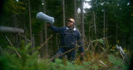 Téléchargez les photos : Un homme caucasien portant un casque enregistre les sons de la nature pour un film en bois de conifères à l'aide d'un équipement audio professionnel. Sound designer travaille avec microphone pare-brise en fourrure à l'extérieur. Cinéma. - en image libre de droit