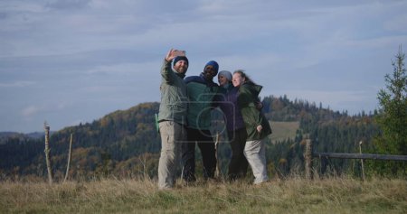 Foto de Grupo de viajeros multiétnicos tomar selfie en el teléfono frente a los paisajes naturales de pie en la cima de la colina. Amigos felices o amigos de senderismo durante las vacaciones en las montañas. Recreación activa. - Imagen libre de derechos