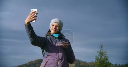 Photo for Female hiker takes selfie using mobile phone standing in front of beautiful scenery on the hill. Caucasian woman holds cup of tea and looks at smartphone. Tourist during trip or trek in the mountains. - Royalty Free Image