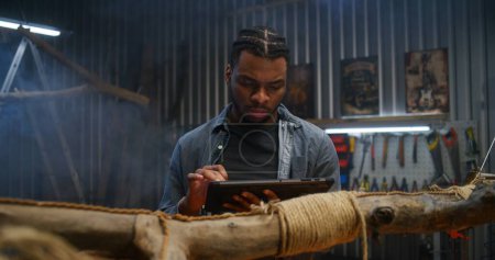 Téléchargez les photos : Créatif concepteur afro-américain utilise tablette numérique tout en créant des produits en bois design dans le garage moderne. Artisanat ou menuisier travaille dans un atelier élégant. Fait main et entrepreneuriat. - en image libre de droit