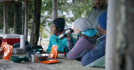 Photo for Tourist friends heat hiking food in pouches and rest in gazebo after long walking expedition in the forest. Group of diverse tourists laugh and talk during trip in the mountains. Active recreation. - Royalty Free Image
