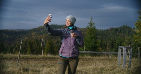 Foto de Senderista toma selfie utilizando el teléfono móvil de pie delante de hermosos paisajes en la colina. Mujer caucásica sostiene la taza de té y mira el teléfono inteligente. Turista durante el viaje o trekking en las montañas. - Imagen libre de derechos