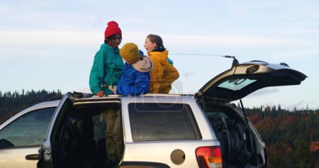 Photo for Young multiethnic tourists sit on the car roof, then get out and unpack luggage and backpacks from the trunk. Group of travelers on vacation road trip in mountains. Outdoor enthusiasts. Slow motion. - Royalty Free Image