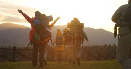 Photo for Diverse hikers with backpacks and trekking poles go hiking in mountains. Happy tourist family or hiking buddies during vacation road trip. Sunset and mountain landscapes in background. Slow motion. - Royalty Free Image