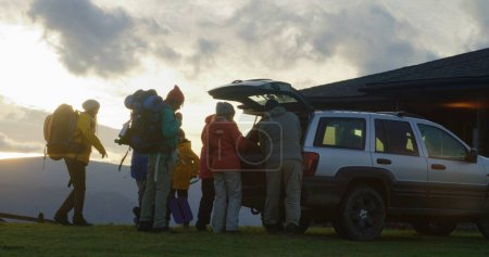 Photo for Full shot of diverse tourist friends arrive to rental house on hill. Happy hikers take luggage from trunk of car and talk. Hiking buddies during trip or expedition to mountains on their vacation. - Royalty Free Image
