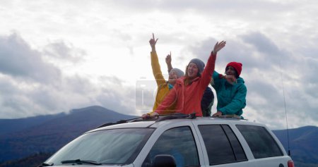 Foto de Grupo de diversos turistas viajan juntos de vacaciones en las montañas. Los excursionistas de pie a través del techo corredizo del coche cantan, bailan y saludan con las manos en el viaje por carretera. A la gente le gusta conducir en la escotilla. Recreación activa. - Imagen libre de derechos