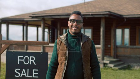 Téléchargez les photos : L'agent immobilier afro-américain se tient près du panneau de vente, regarde la caméra et sourit. Homme agent immobilier attend les visiteurs de la maison élégante moderne dans les montagnes. Propriété résidentielle en vente concept. - en image libre de droit
