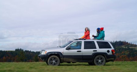 Photo for Group of diverse tourists together ride on vacation in mountains. Hikers standing up through sunroof of car sing, dance and wave hands on road trip. People enjoy drive in car hatch. Active recreation. - Royalty Free Image