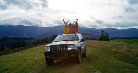 Foto de Grupo de turistas multiétnicos viajan juntos de vacaciones en las montañas. Los excursionistas de pie a través del techo corredizo del coche, reír y agitar las manos en el viaje por carretera. Disfrutando de conducir en una escotilla de coche. Entusiastas al aire libre. - Imagen libre de derechos