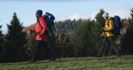 Foto de Foto completa de pareja multiétnica de turistas caminando a lo largo del sendero en el bosque en otoño. Hombre afroamericano y mujer caucásica durante el viaje o caminata a las montañas. Turismo y exploración al aire libre. - Imagen libre de derechos