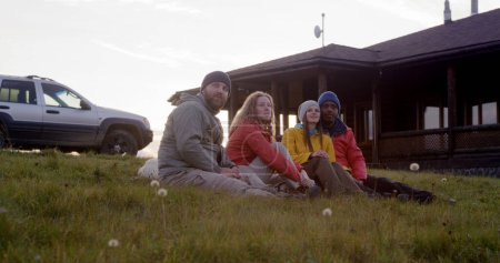 Foto de Grupo de turistas multiétnicos se sientan en la hierba en la cima de la colina y hablan. Los viajeros o amigos de senderismo disfrutan de la naturaleza durante el viaje a las montañas. Elegante casa de madera, coche y perro en el fondo. - Imagen libre de derechos
