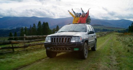 Foto de Grupo de turistas multiétnicos viajan juntos de vacaciones en las montañas. Los excursionistas de pie a través del techo corredizo del coche, reír y agitar las manos en el viaje por carretera. Disfrutando de conducir en una escotilla de coche. Entusiastas al aire libre. - Imagen libre de derechos