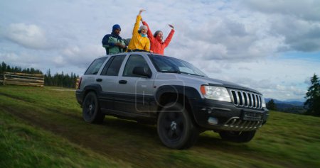 Foto de Grupo de turistas multiétnicos viajan juntos de vacaciones en las montañas. Los excursionistas de pie a través del techo corredizo del coche, reír y agitar las manos en el viaje por carretera. A la gente le gusta conducir en la escotilla. Entusiastas al aire libre - Imagen libre de derechos