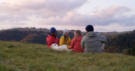 Foto de Grupo de turistas multiétnicos se sientan en la hierba en la cima de la colina y acarician a un perro. Los viajeros o amigos de senderismo disfrutan de la naturaleza durante el viaje. Puesta de sol y hermoso paisaje de montaña. Concepto de recreación activa. - Imagen libre de derechos