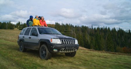 Foto de Grupo de turistas multiétnicos viajan juntos de vacaciones en las montañas. Los excursionistas de pie a través del techo corredizo del coche, reír y agitar las manos en el viaje por carretera. A la gente le gusta conducir en la escotilla. Entusiastas al aire libre - Imagen libre de derechos
