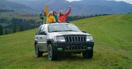Photo for Group of diverse outdoor enthusiasts together ride on vacation in mountains. Hikers standing up through sunroof of car sing, dance and wave hands on road trip. Hiking buddies enjoy drive in car hatch. - Royalty Free Image