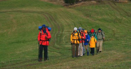 Photo for Full shot of diverse tourist friends with backpacks and trekking poles walking along trail on hill in camp. Hikers during trip or expedition to mountains on vacation to mountains. Tourism and travel. - Royalty Free Image